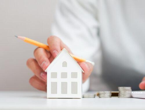 Mann stellt Berechnungen für Immobilienfinanzierung an, vor ihm ein Münzstapel und ein Modell eines Hauses
